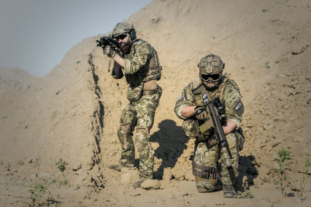 Two soldiers in camouflage gear equipped with weapons in a desert environment.